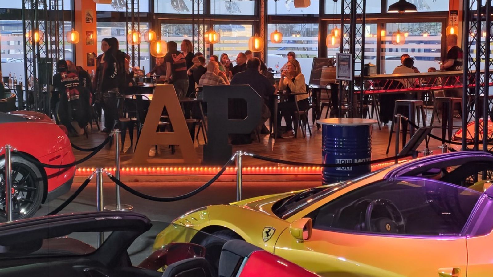 Restaurantbereich im AP Café INDOOR-RACING Aadorf: besetzte Tische unter warmen Hängelampen, im Vordergrund ausgestellte Ferrari-Sportwagen hinter einer Absperrung, in der Mitte der grosse AP-Schriftzug