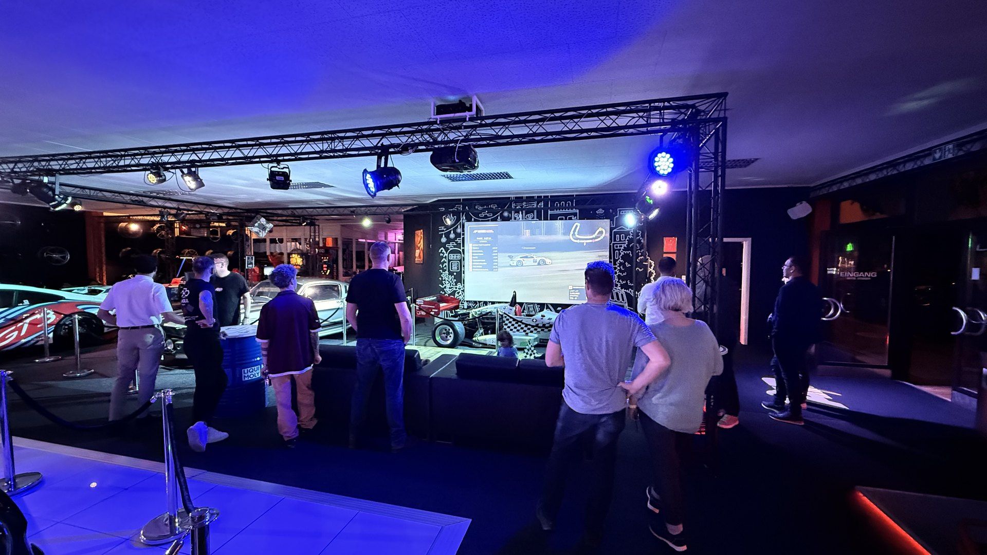 Public Viewing im Sim Racing Center Aadorf: Gäste verfolgen das Rennen auf der grossen Leinwand, daneben Sport- und Rennwagen der Auto-Ausstellung