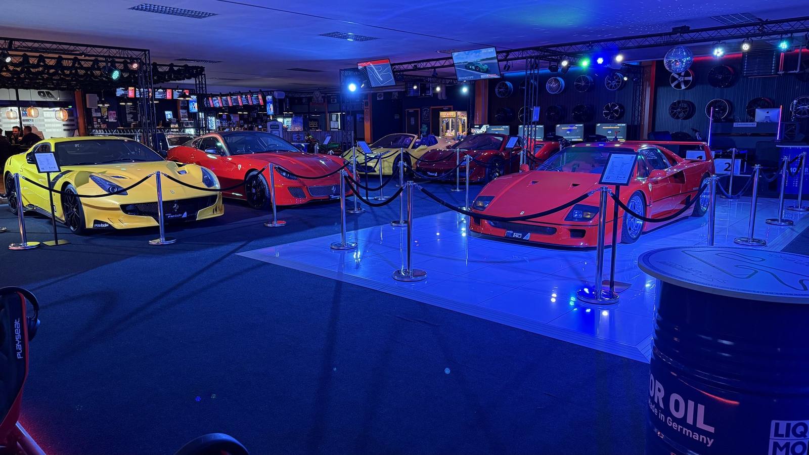 Auto-Ausstellung im Sim Racing Center Aadorf: Ferrari F40, F12 tdf und weitere Sportwagen unter blauem Licht, dahinter die Rennsimulatoren im AP Café INDOOR-RACING