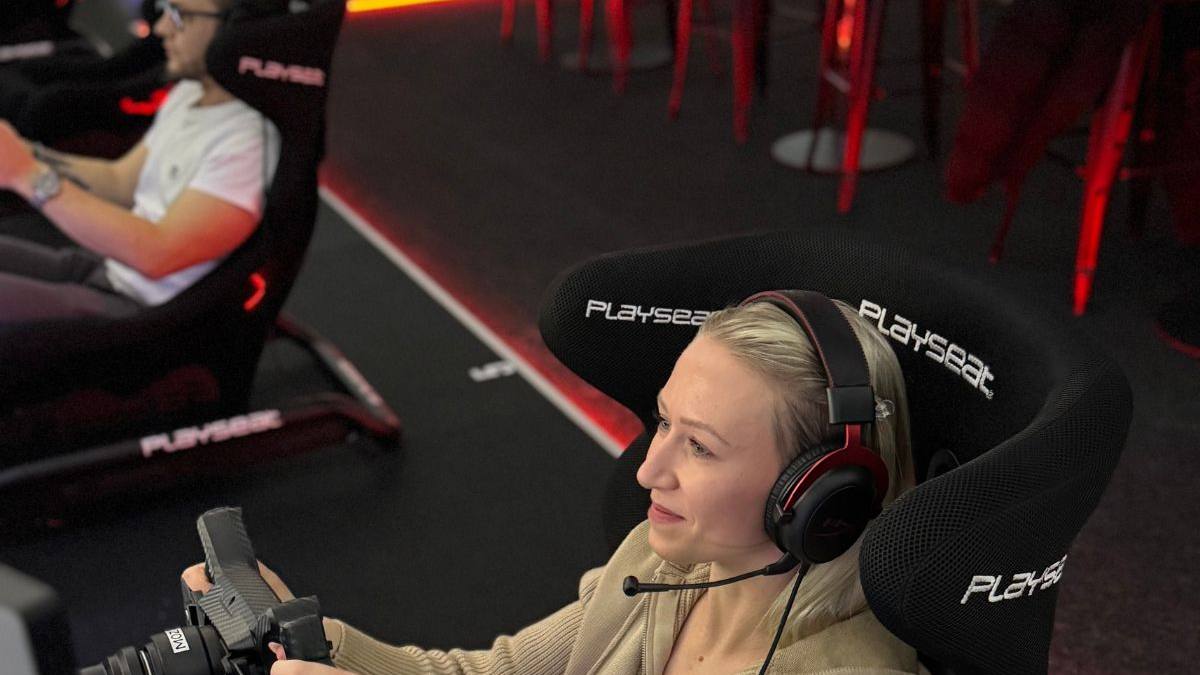 Mitarbeiterin konzentriert am Lenkrad eines Rennsimulators, Kollege im Hintergrund — Sim Racing mit der Firma
