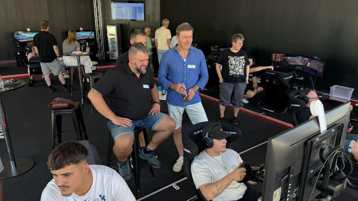 Firmen-Team-Event im Sim Racing Center Aadorf: Teilnehmende mit Namensschildern rund um die Playseat-Rennsimulatoren, ein Fahrer mit Headset am Lenkrad vor dem gebogenen Bildschirm, im Hintergrund die Anzeigetafel mit den Rundenzeiten