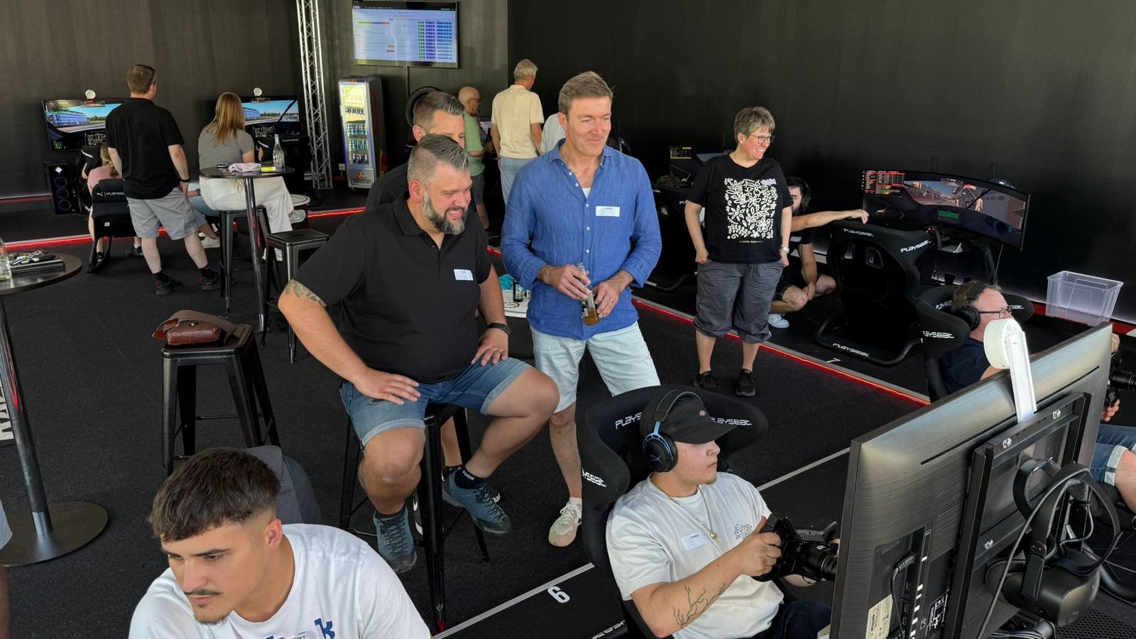 Firmen-Team-Event im Sim Racing Center Aadorf: Teilnehmende mit Namensschildern rund um die Playseat-Rennsimulatoren, ein Fahrer mit Headset am Lenkrad vor dem gebogenen Bildschirm, im Hintergrund die Anzeigetafel mit den Rundenzeiten
