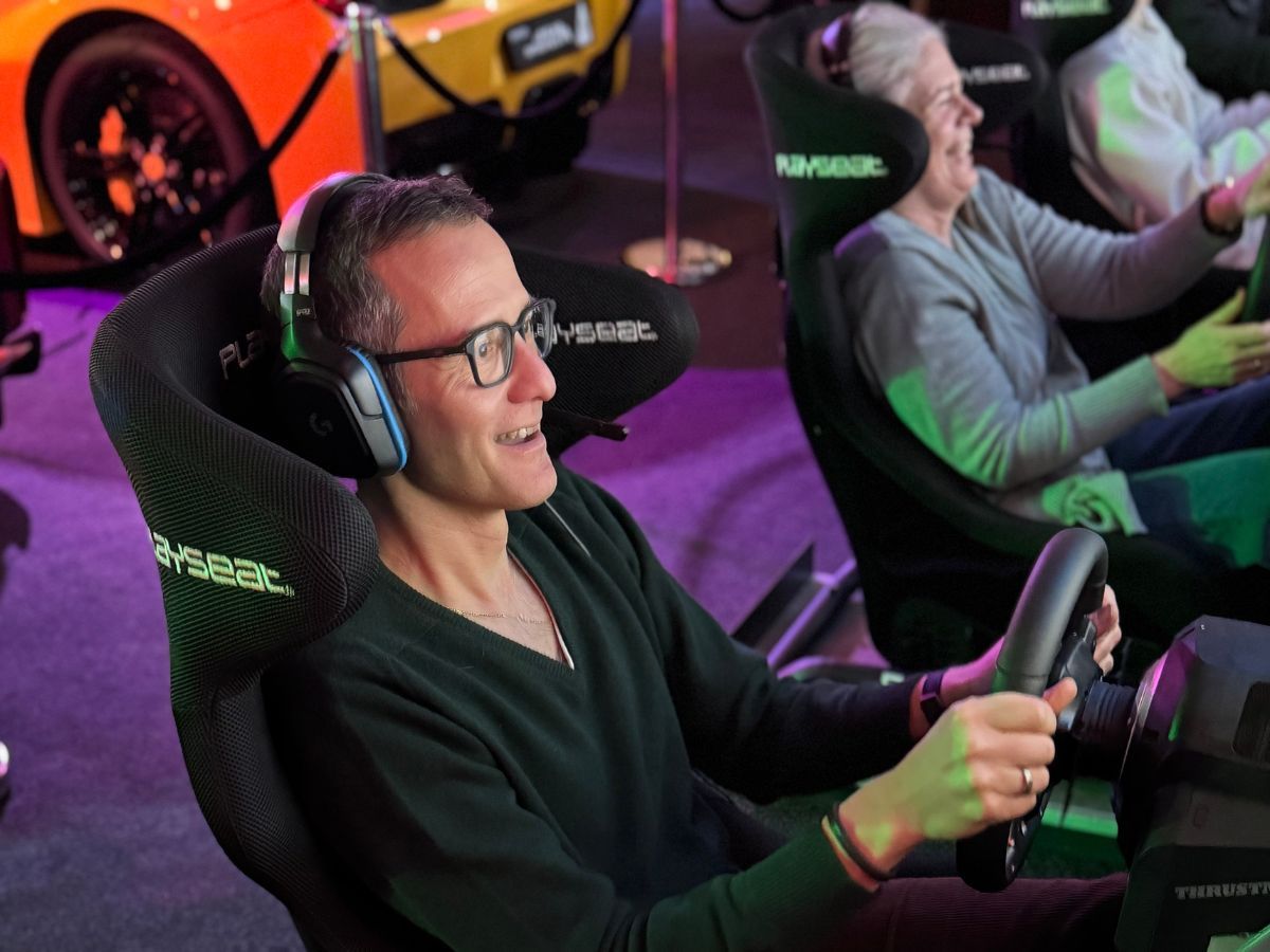 Fahrer und ältere Frau lachen am Lenkrad, im Hintergrund ein Sportwagen — Spass beim Sim Racing