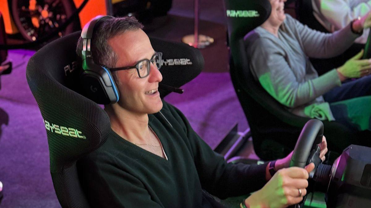 Fahrer und ältere Frau lachen am Lenkrad, im Hintergrund ein Sportwagen — Spass beim Sim Racing