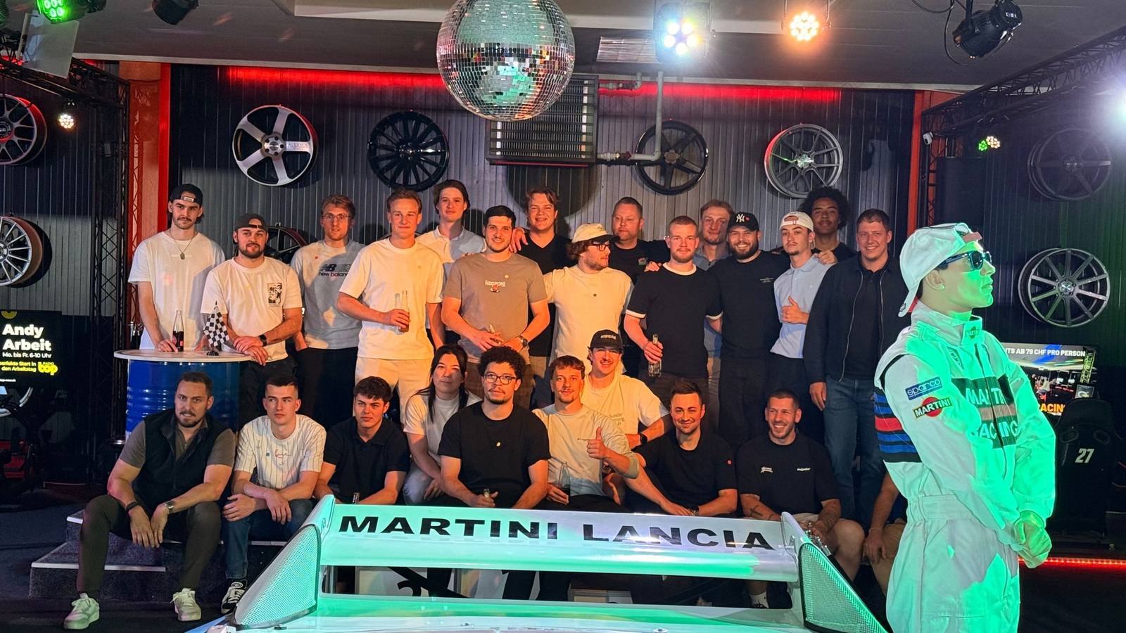Grosse Gruppe beim Gruppenfoto vor dem Martini-Lancia-Rennwagen und der Felgenwand im Sim Racing Center Aadorf — Team-Event aus der Region Wil