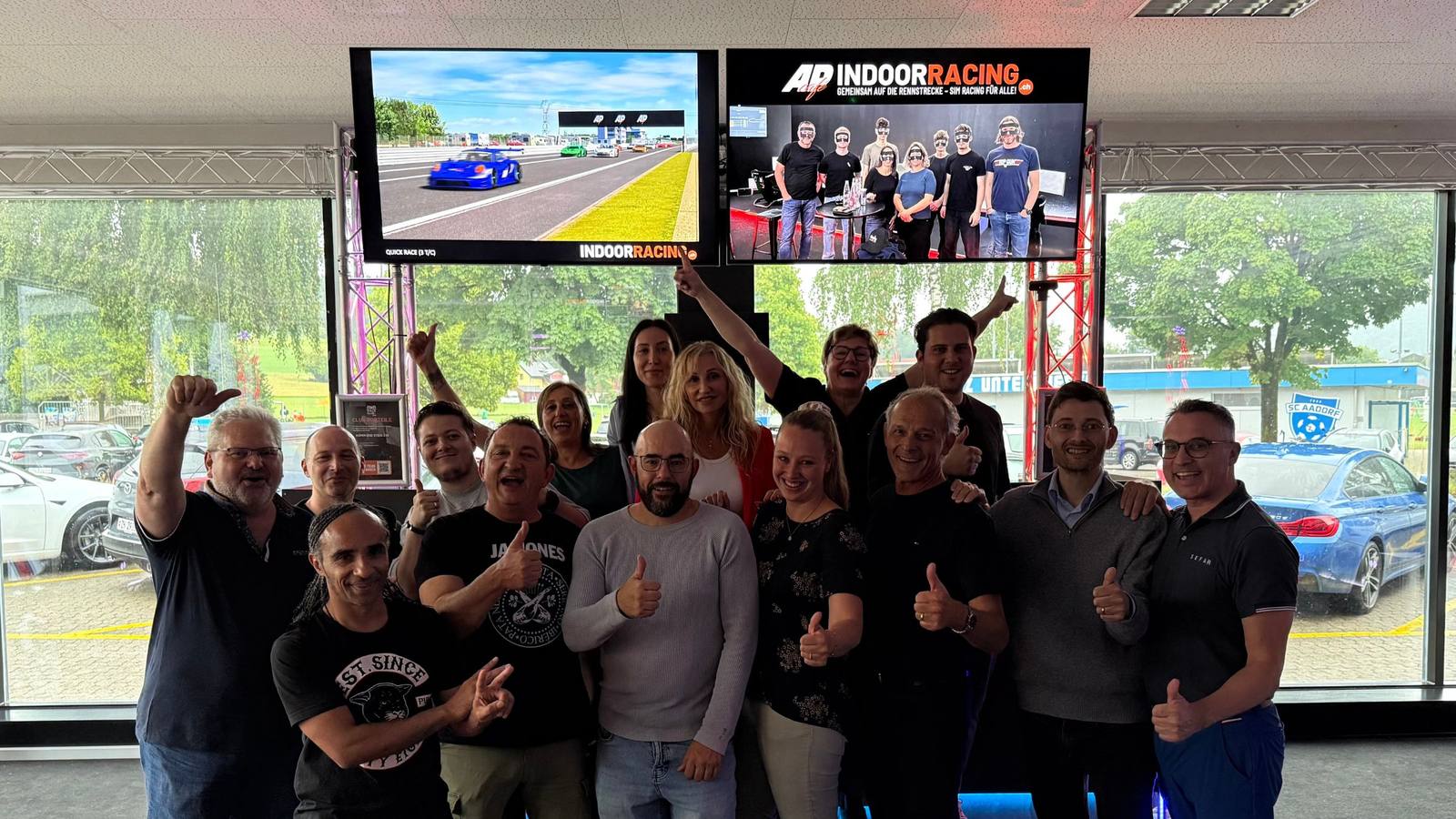 Firmenteam jubelt mit Daumen hoch vor den Rennbildschirmen im Sim Racing Center Aadorf, auf dem Banner steht „AP Café INDOOR RACING — gemeinsam auf die Rennstrecke, Sim Racing für alle!" — Team-Event einer Firma aus Winterthur