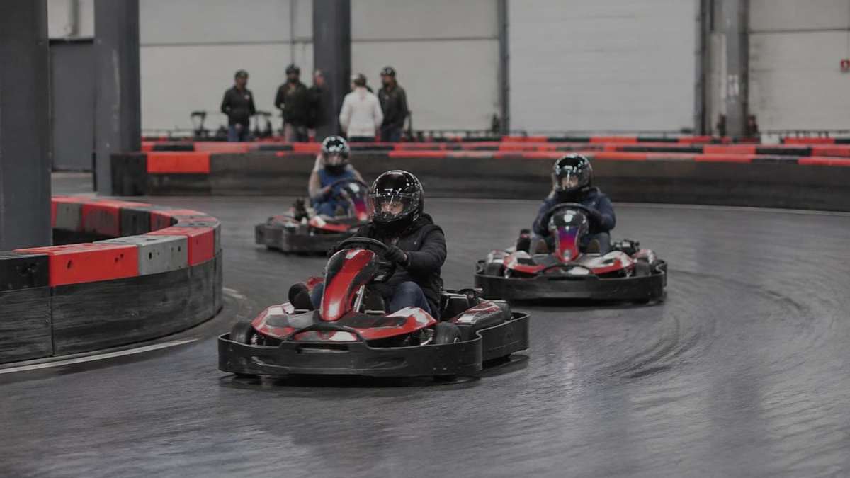 Go-Kart-Fahrer mit Helmen in einer Kurve, eine Gruppe wartet an der Bande (KI-Symbolbild)