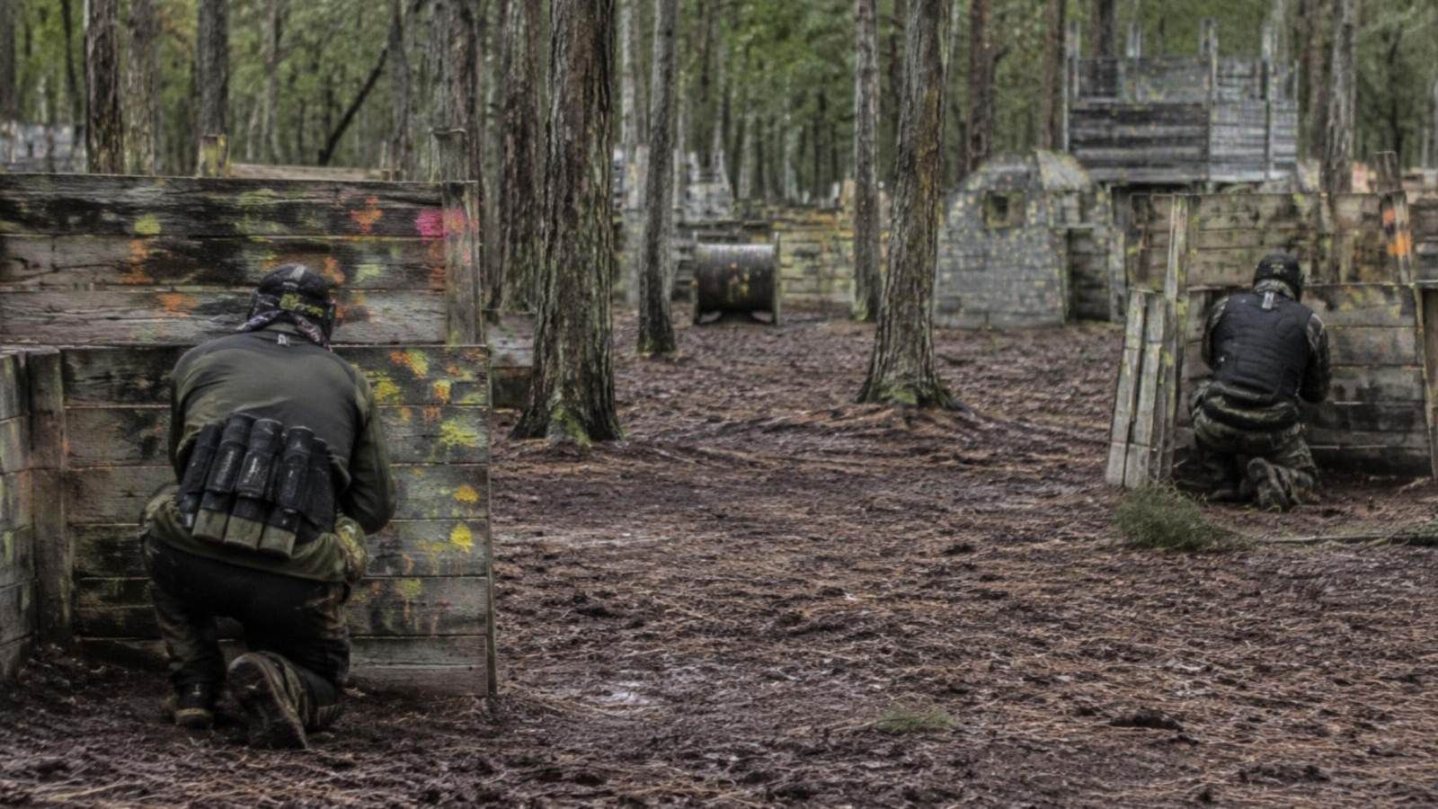 Paintball-Spieler von hinten hinter Holzbarrikaden mit Farbspritzern im Wald (KI-Symbolbild)