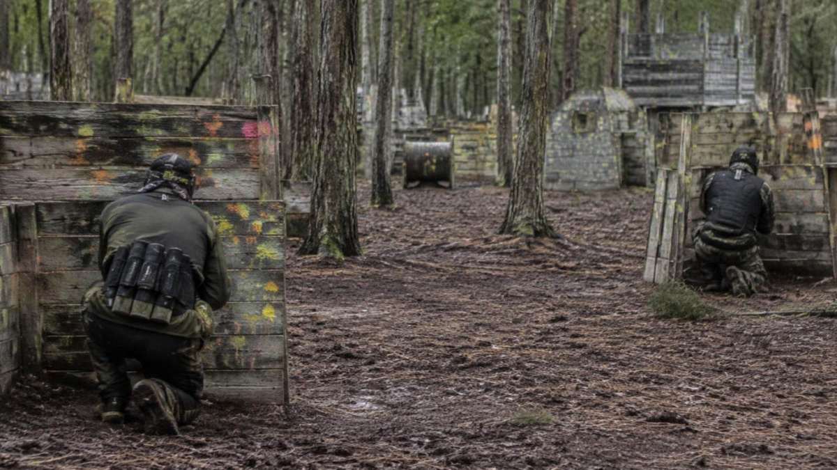 Paintball-Spieler von hinten hinter Holzbarrikaden mit Farbspritzern im Wald (KI-Symbolbild)